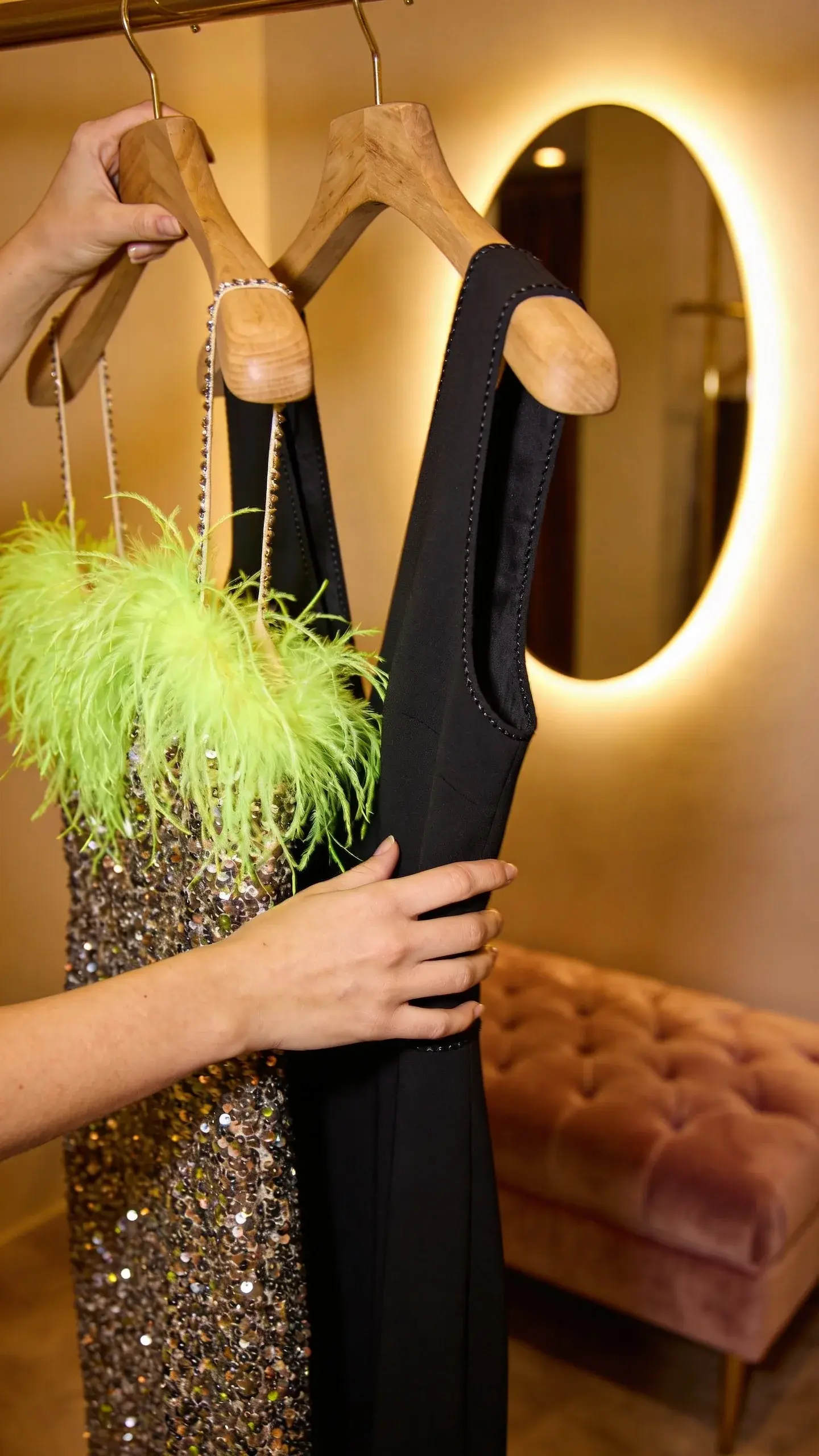 A softly lit boutique fitting room: close-up of hands choosing between a sequin-studded, neon feather-trimmed dress and a simple, impeccably cut black column dress on wooden hangers; focus on textures and stitching, velvet bench and mirror glow, no faces visible.