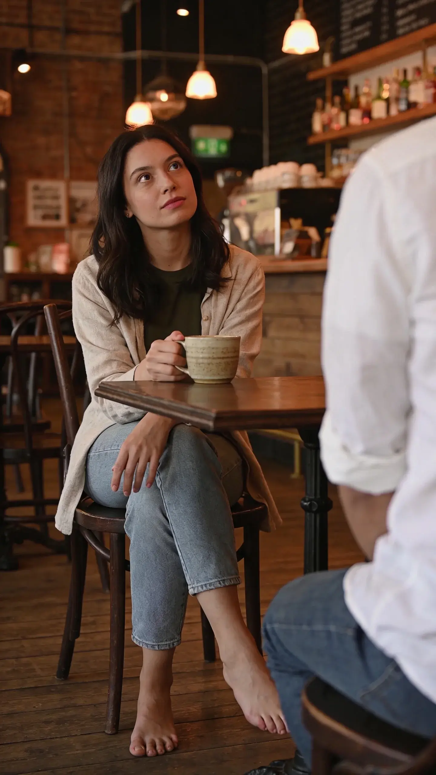 Over-the-shoulder cinematic shot in a café: a woman seated with feet grounded, ankles uncrossed, spine straight but relaxed; one hand loosely around a ceramic cup, the other resting still on her knee; gaze lifted toward someone just off-frame, steady and attentive; warm, ambient light, wood textures, muted tones; no fidgeting props.