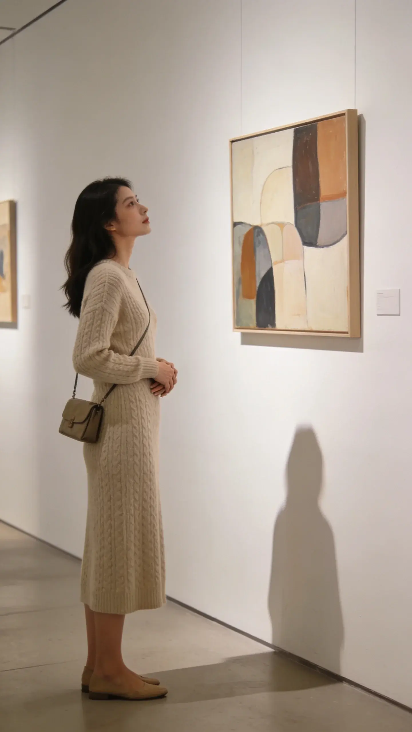 Gallery interior: a woman stands before abstract art, feet firmly planted, spine tall; she leans in slightly with composed curiosity; hands loosely clasped at her waist, no restless movements; elegant knit dress, small crossbody bag; soft museum lighting, white walls, subtle shadows.