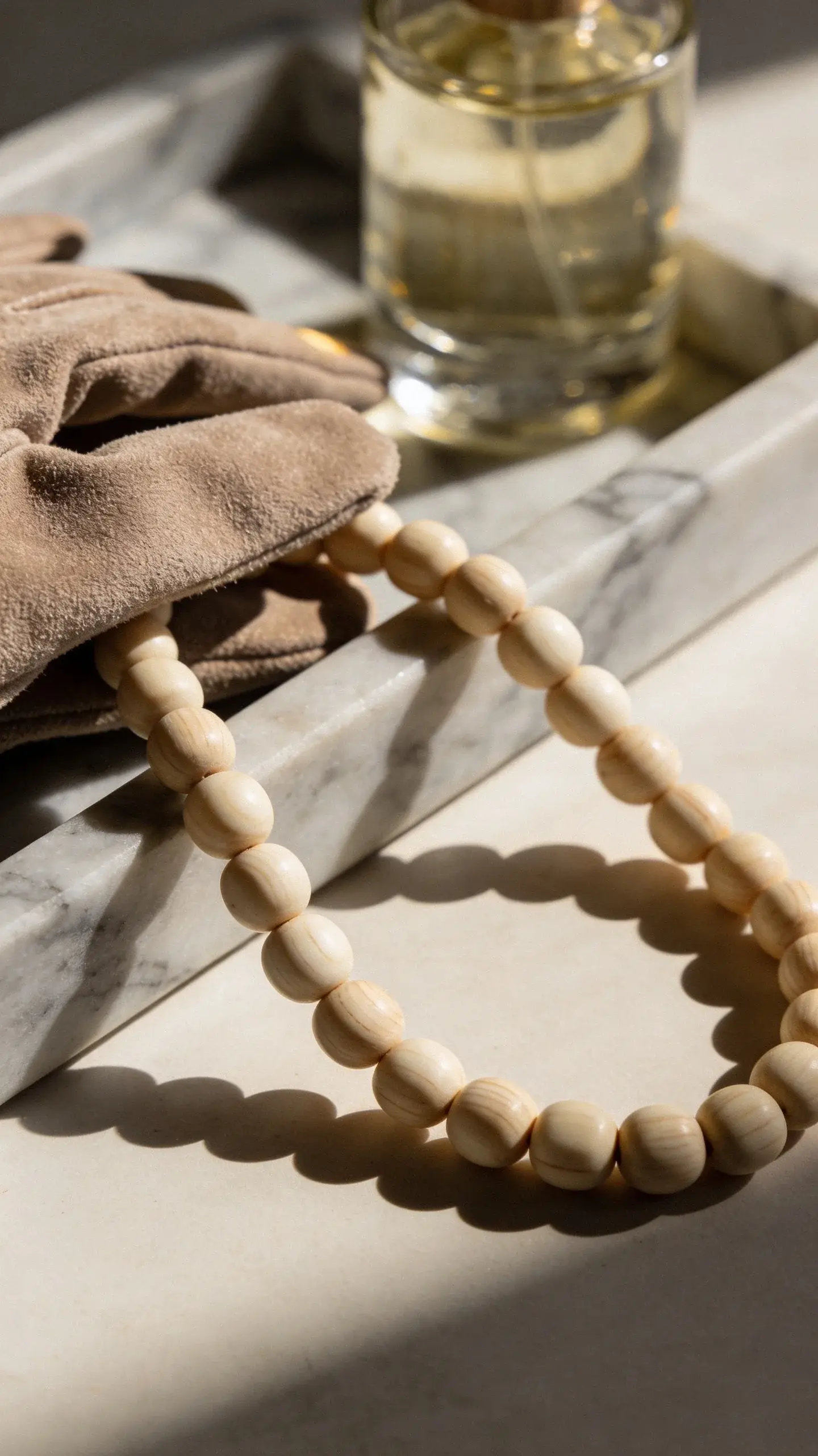 Close-up of creamy sandalwood beads and a suede glove in taupe draped over a marble tray; a thin ribbon of sunlight creates gentle highlights; an unfocused bottle of translucent perfume in the background; tones of beige, cream, and warm gray; tactile textures emphasized; no branding or text.