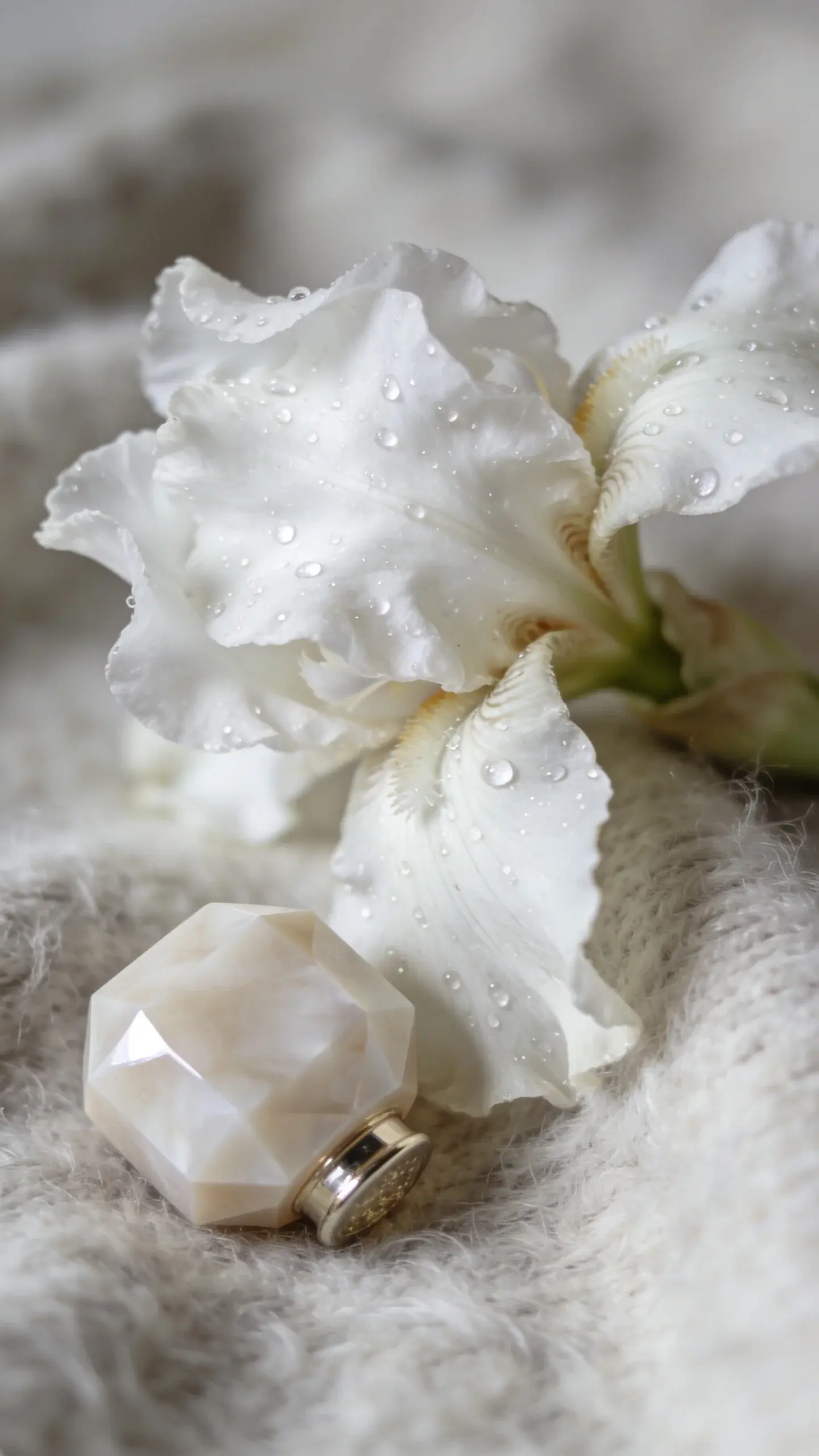 Close-up macro shot of white iris petals against a backdrop of brushed cashmere; dew-like droplets glinting softly; a pale, faceted perfume stopper lies beside; creamy whites and pale greys with delicate bokeh; ethereal, quiet mood; no text.