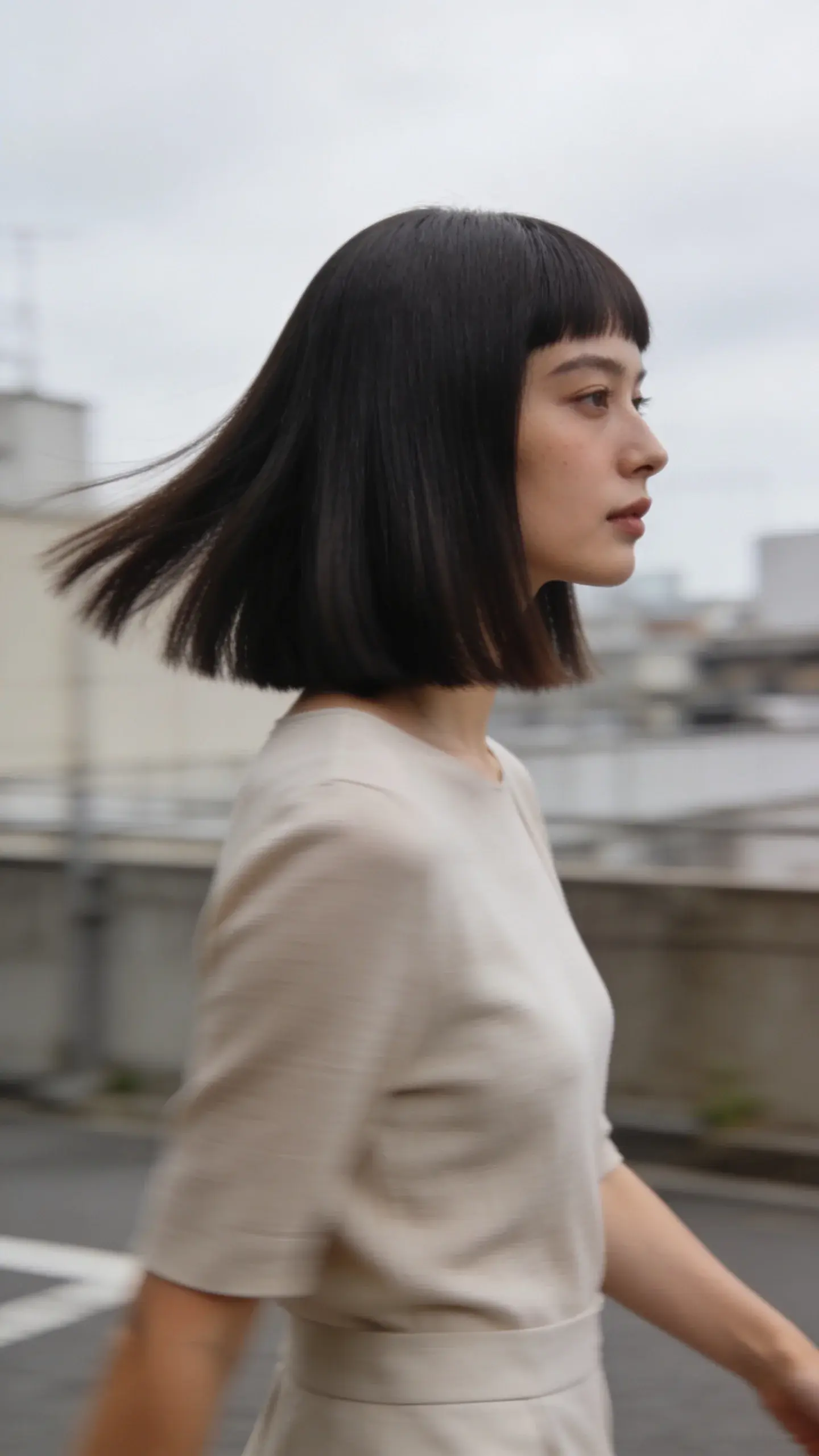 Candid-style image of a woman walking outdoors with a sleek, straight, one-length cut just past the shoulders; movement without frizz, ends perfectly even; soft overcast daylight creating a naturally expensive finish; urban neutral background.