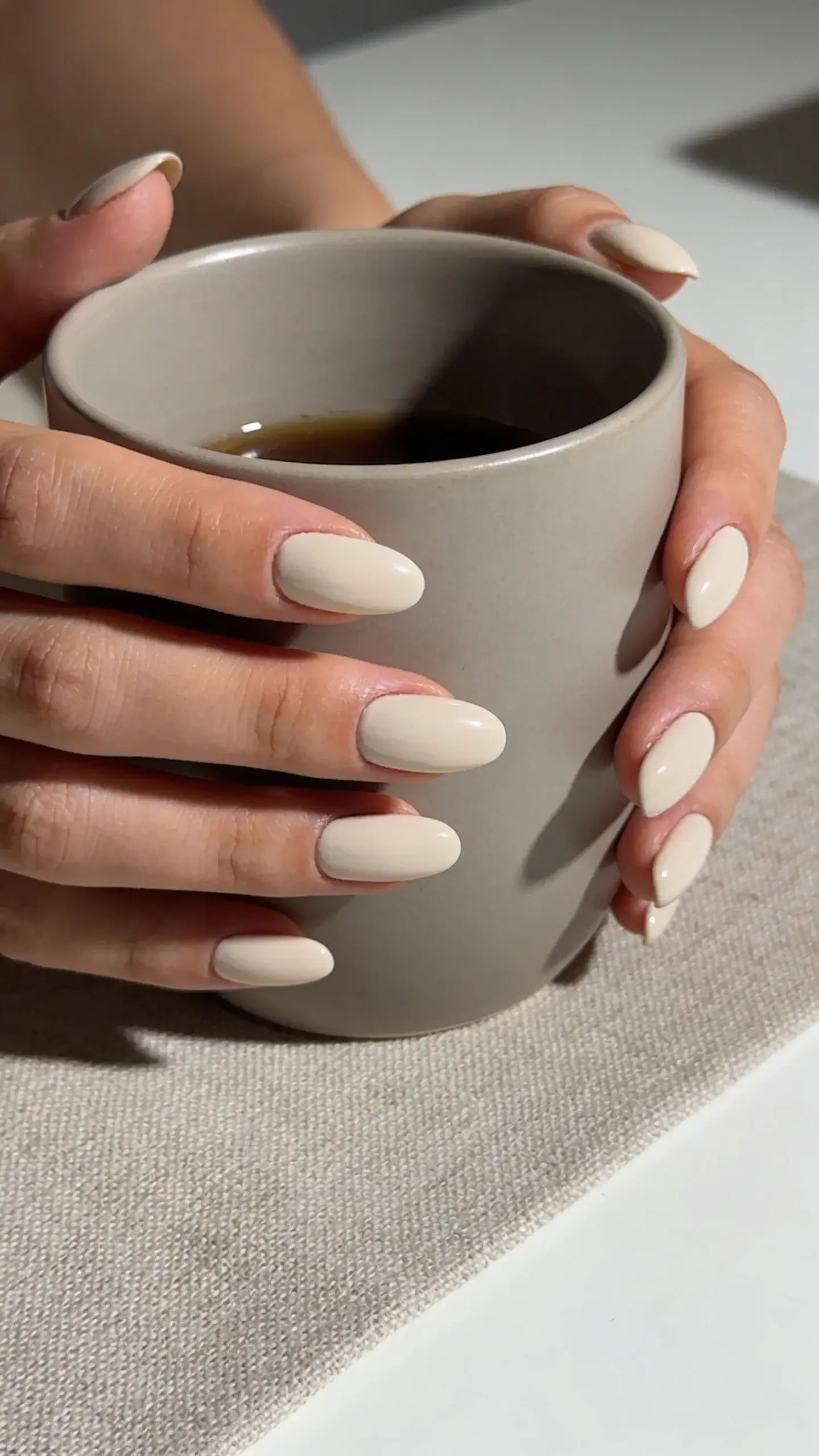 Studio shot of medium-length oval nails painted in a cool beige-taupe cream polish; hands lightly holding a matte ceramic coffee cup in a greige tone; soft shadowing; clean, minimalist desk scene with a linen placemat; no text.