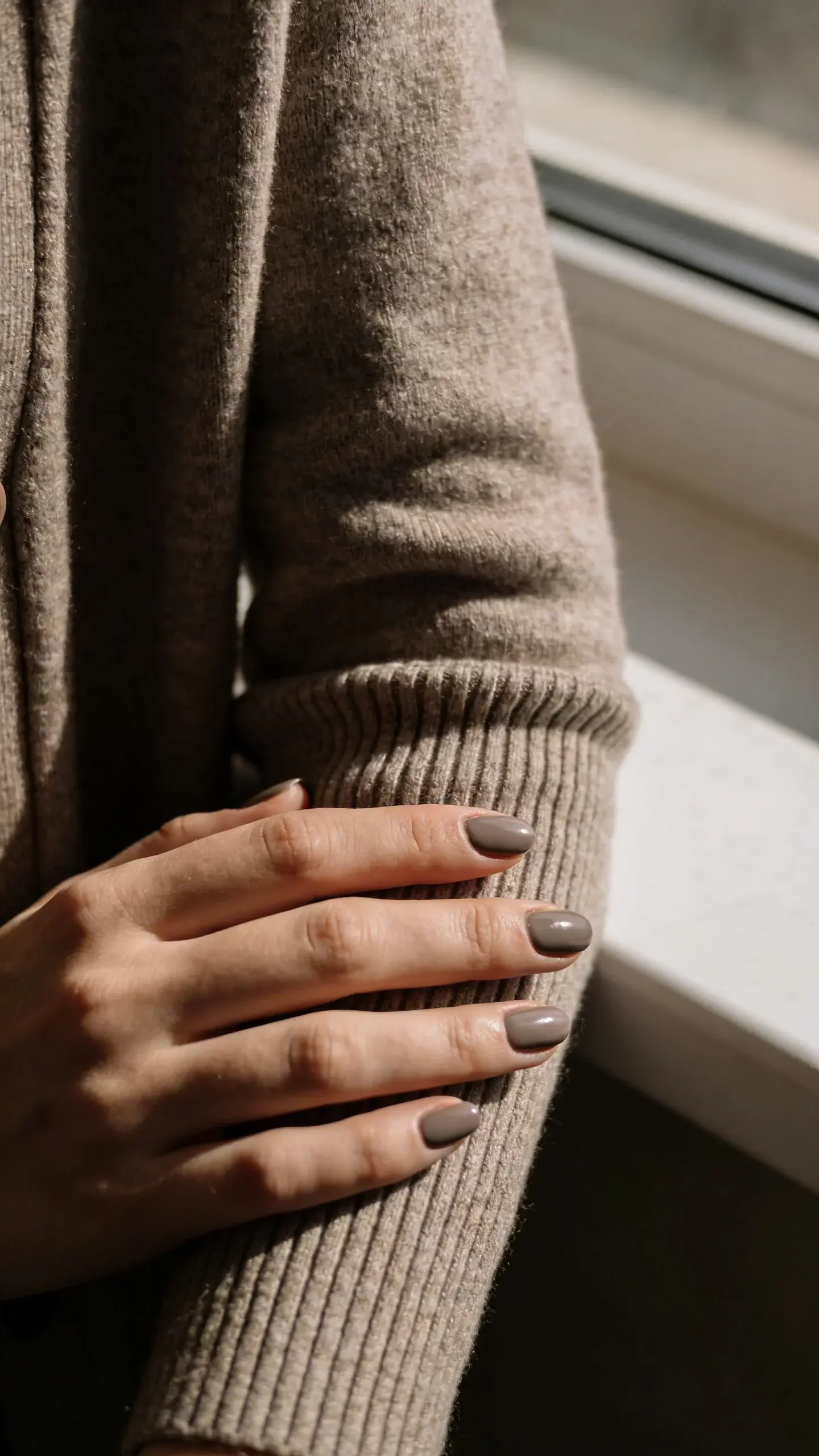 Cozy lifestyle shot of hands with soft mushroom-gray nails (cool undertone) resting on a ribbed cashmere cardigan sleeve; warm window light; subtle texture of knit fabric in frame; muted, monochrome palette with taupe and mushroom hues; no text.