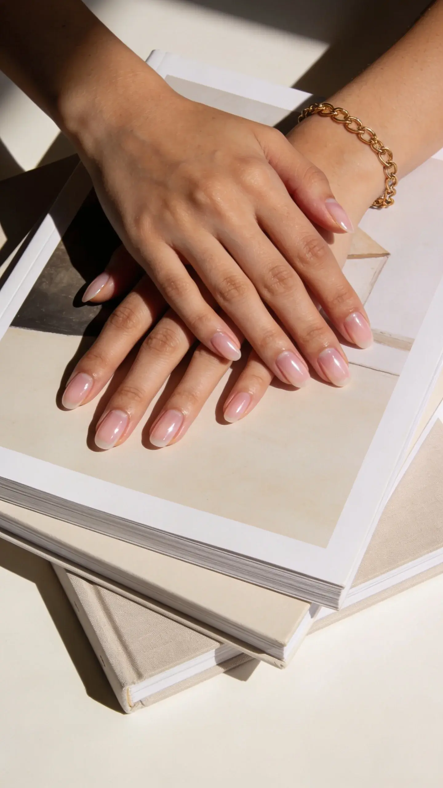 Flat-lay of two hands with sheer milky pink nails, glossy, resting over a stack of art books with neutral covers; delicate gold chain bracelet; background in soft ivory; natural light from left casting gentle shadows; no text.