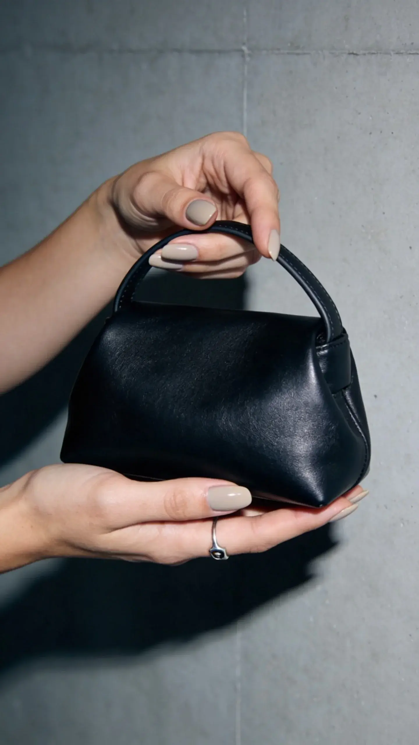 Editorial-style image of hands with beige-taupe nails holding a sleek black leather mini bag; cool-toned lighting; minimal jewelry (thin silver signet ring); backdrop a smooth cement-gray wall; no text.