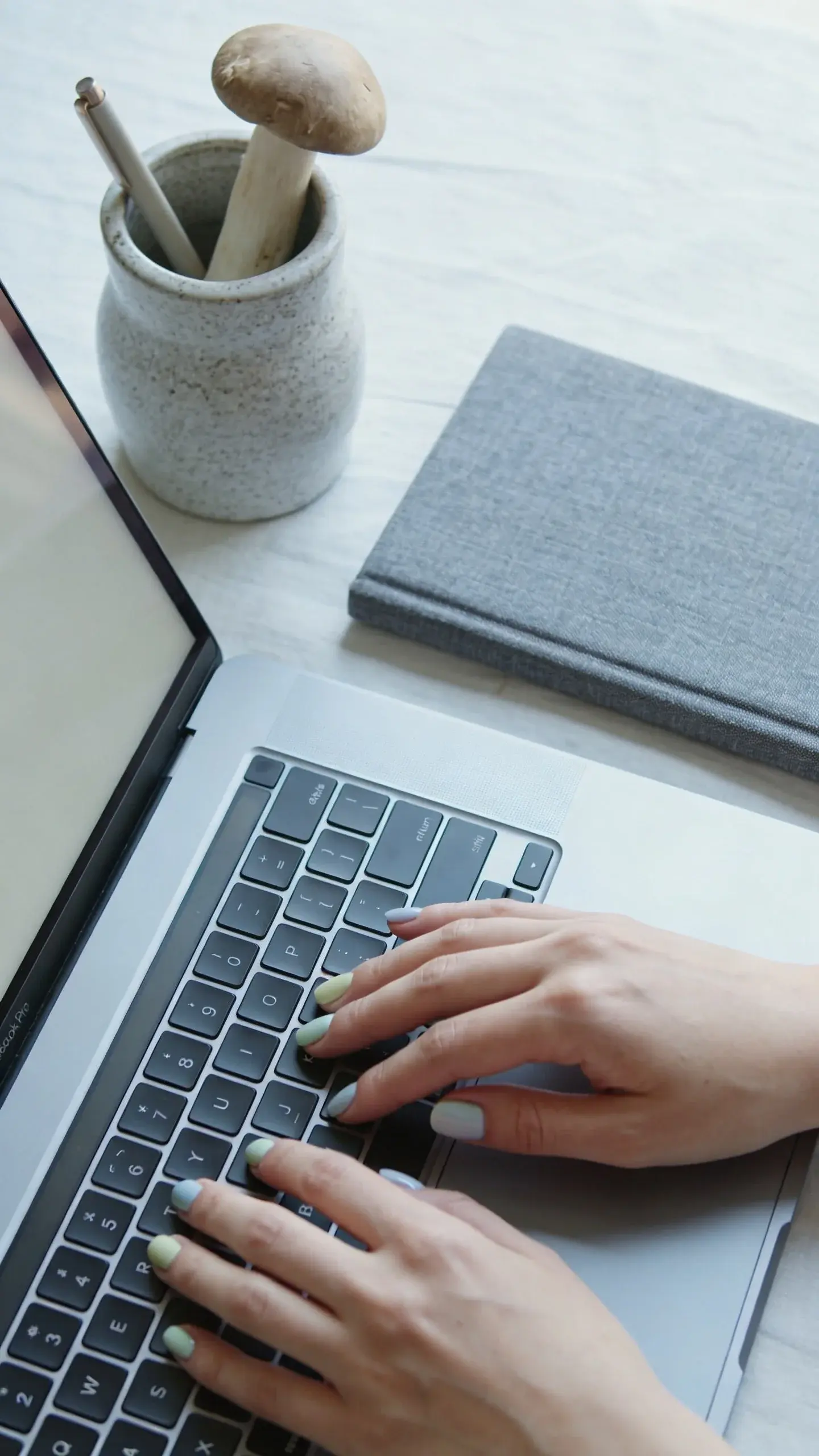 Lifestyle desk scene: hands with soft mushroom polish typing on a silver laptop; cool daylight; accessories include a stoneware pen holder and a gray linen notebook; color palette in mushroom, slate, and ivory; no text.
