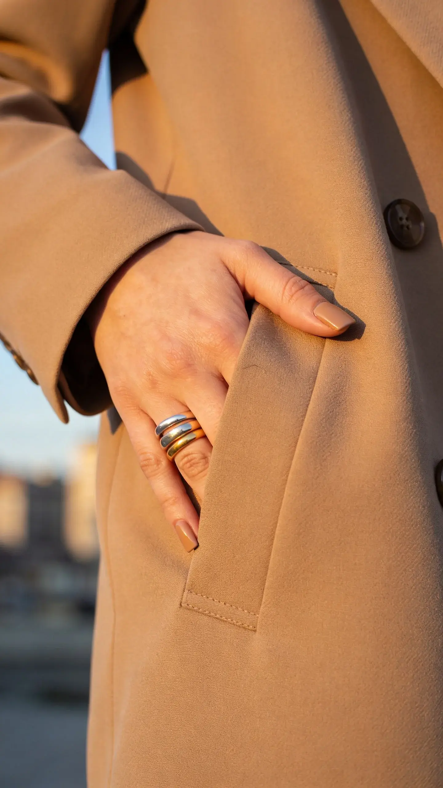 Fashion-forward portrait crop: hand with short square nails in beige-taupe tucked into the pocket of a tailored camel coat; soft golden hour light; blurred city background; ring stack in mixed metals; no text.