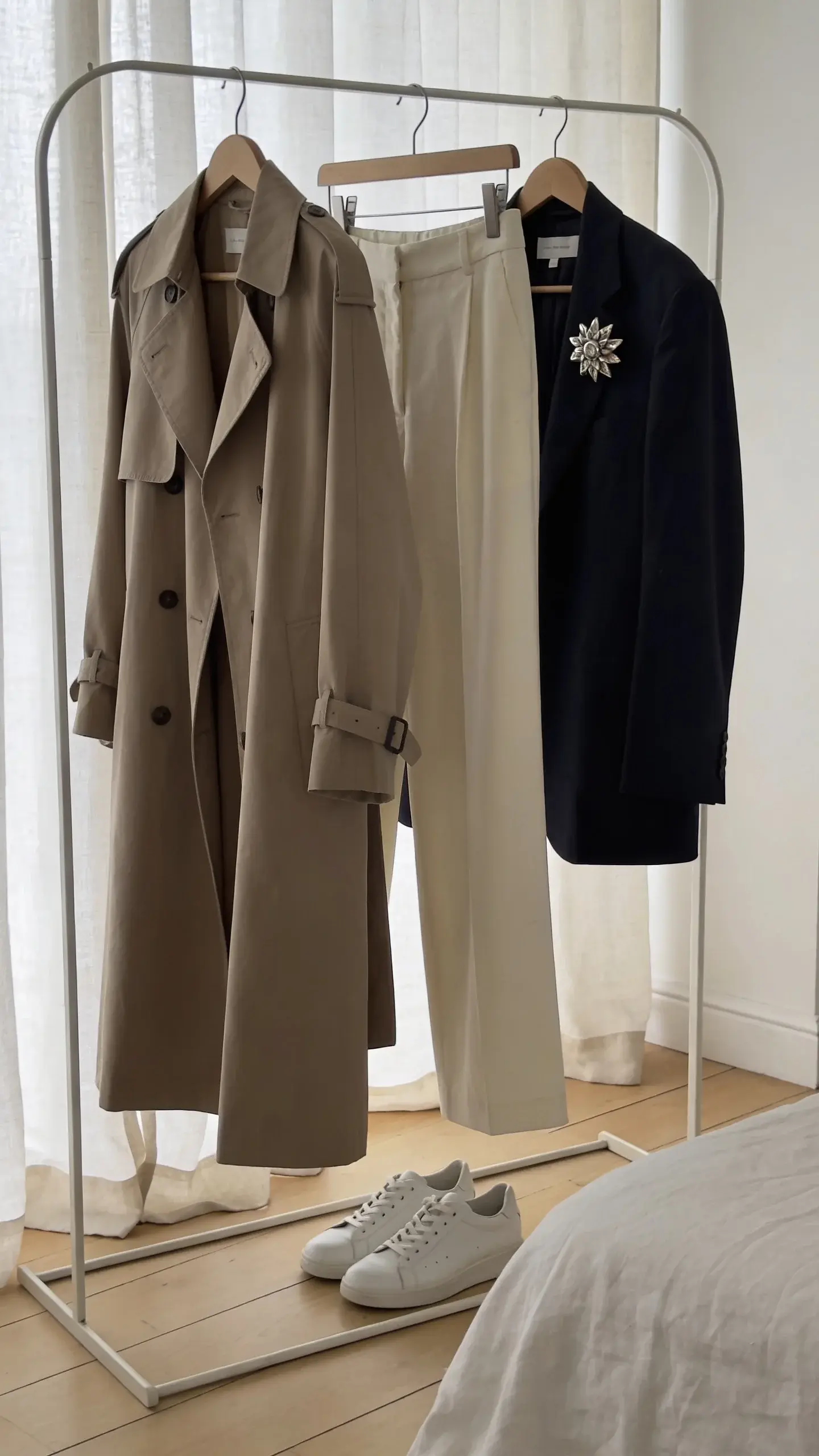 A minimalist bedroom clothing rack with a tight, cohesive capsule: relaxed tailoring pieces in soft neutrals (oatmeal trench, cream wide-leg trousers, navy oversized blazer), clean white leather sneakers, and one eccentric accessory (a single sculptural silver brooch). Natural morning light, pale wood floors, linen curtains, no text, editorial fashion still-life.