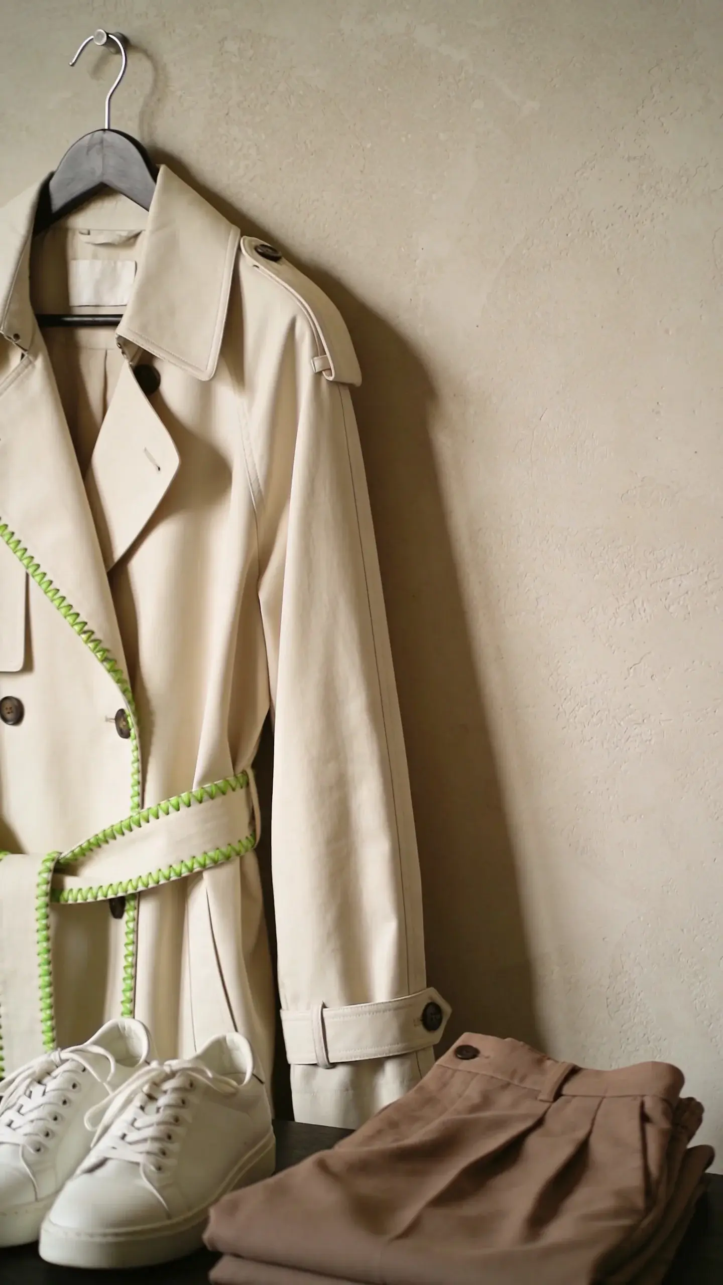 Close-up still-life of a “one weird detail”: cream trench coat on a hanger with a striking, unexpected detail—lime-green contrast stitch along the belt. Nearby: white sneakers, taupe trousers folded. Neutral plaster wall, soft side light, no text.