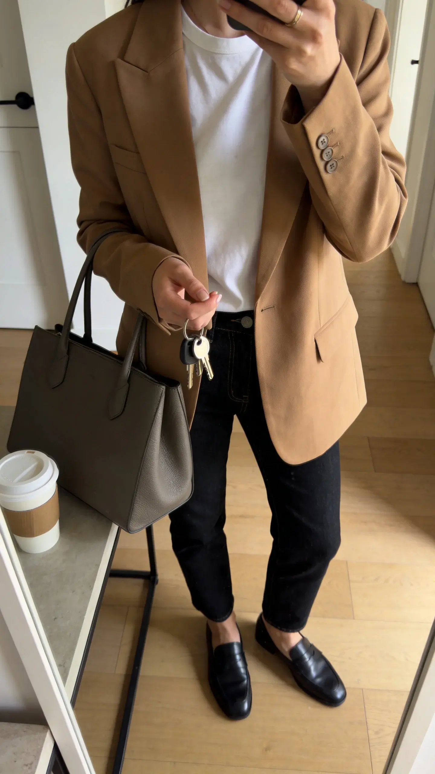 A hallway mirror selfie vibe without showing the face: cropped view of a woman wearing dark jeans, a tucked white tee, a camel blazer, sleek loafers, and a structured tote, keys in hand, coffee on a console—clearly ready for work then errands. Natural morning light, no text.