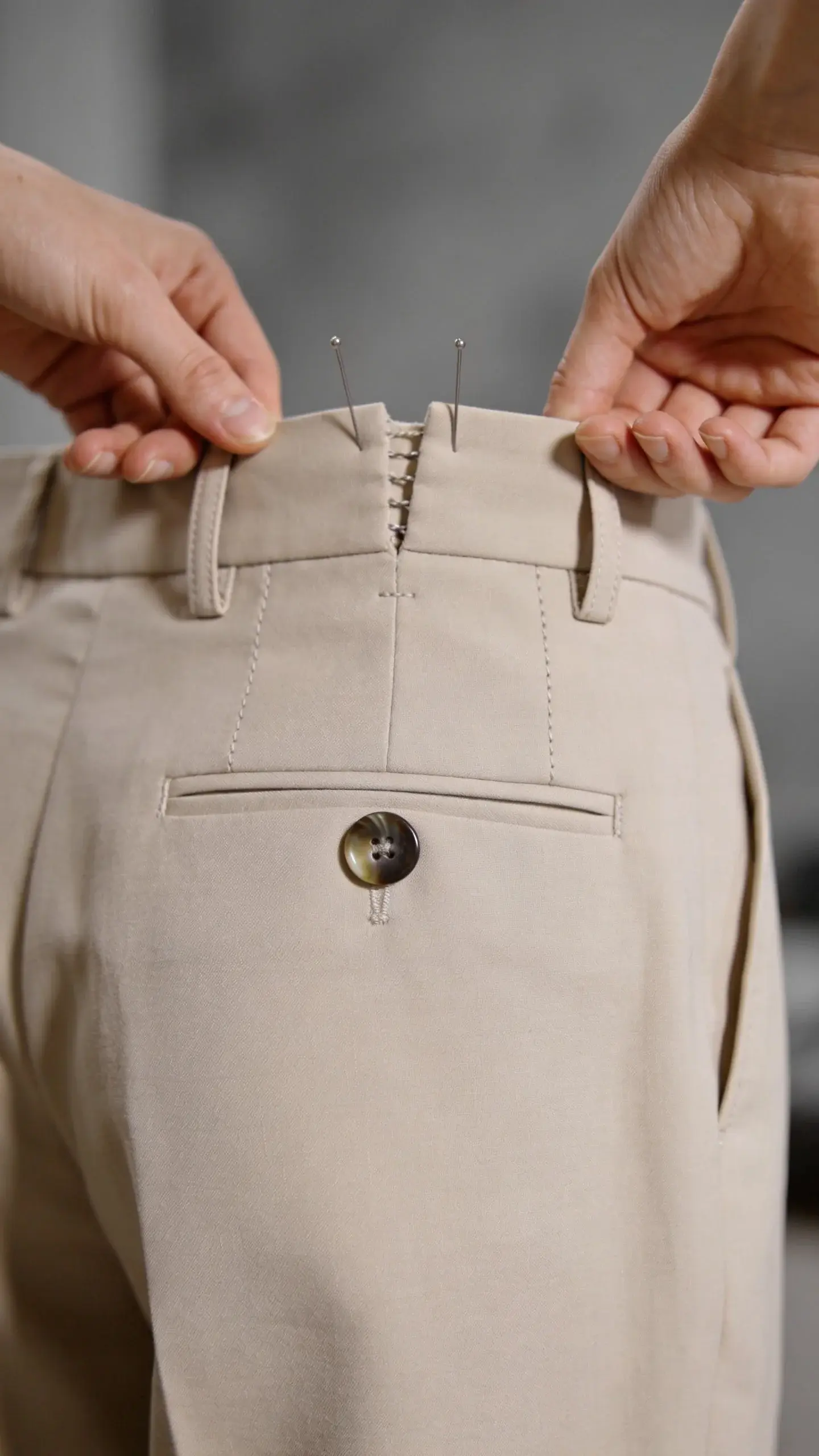 Detail shot of a waistband alteration fitting: hands adjusting the back of high-waisted trousers to remove gaping; two pins holding excess fabric; visible belt loops aligned, neat stitching, matte horn button; neutral beige fabric against a soft gray backdrop, shallow depth of field, no text.